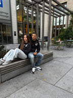 Two models wearing black oversized Clover at Night hoodies by R3CEIVED sitting outdoors on a bench in an urban setting