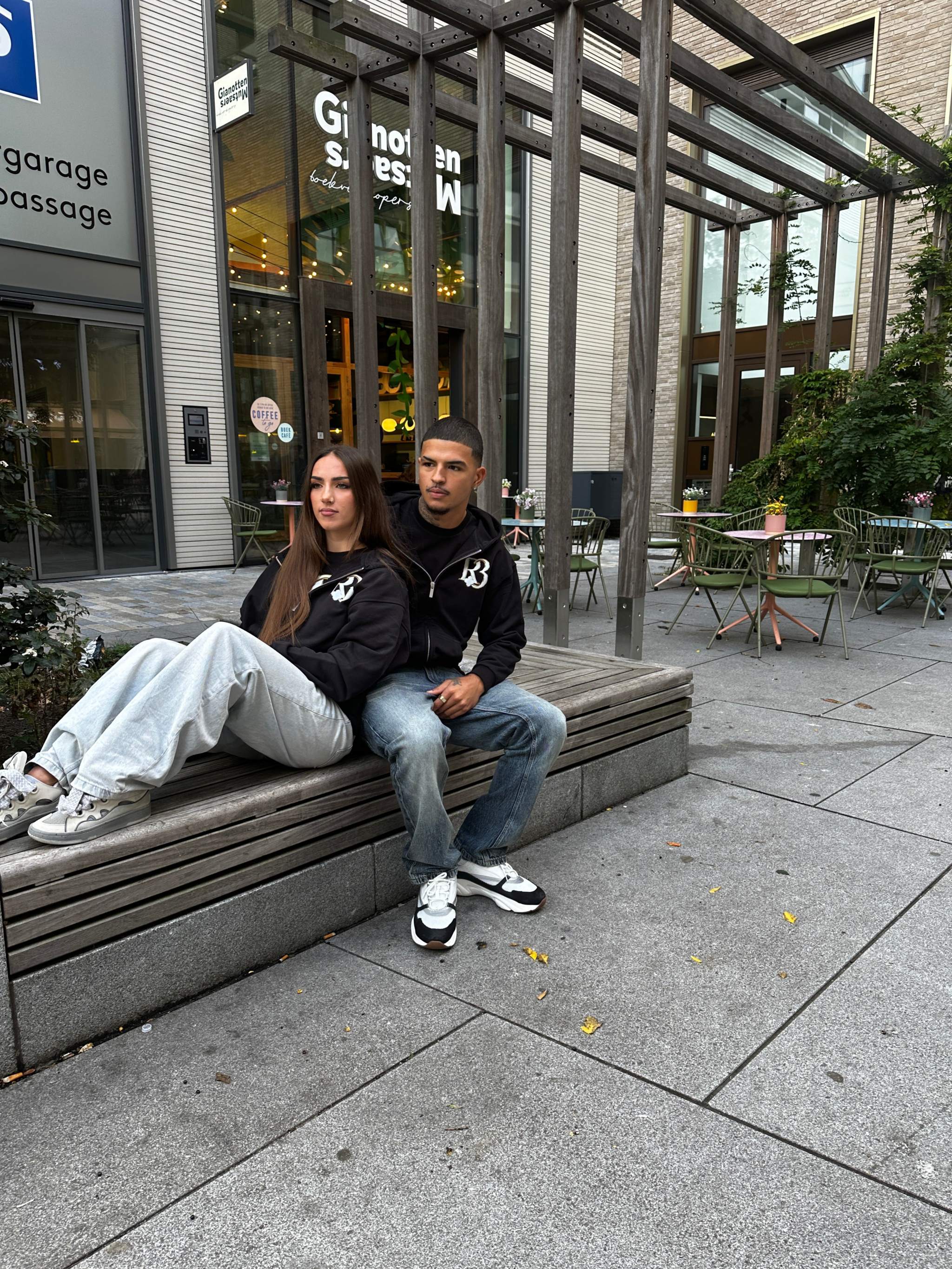 Two models wearing black oversized Clover at Night hoodies by R3CEIVED sitting outdoors on a bench in an urban setting