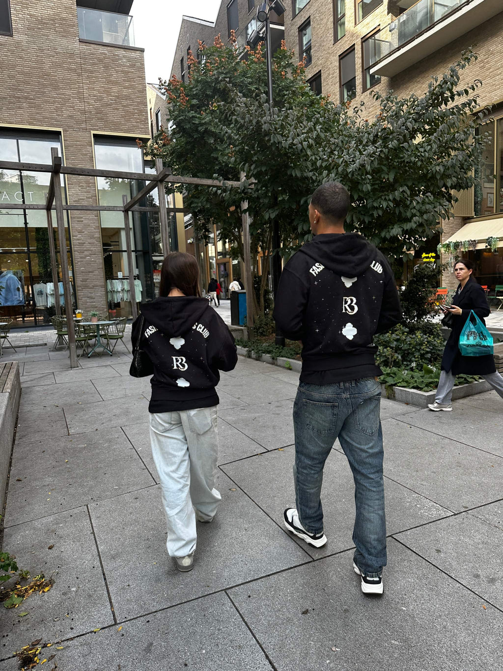 Two people wearing black Clover at Night hoodies with R3 logo and clover designs on the back walking outdoors in an urban setting