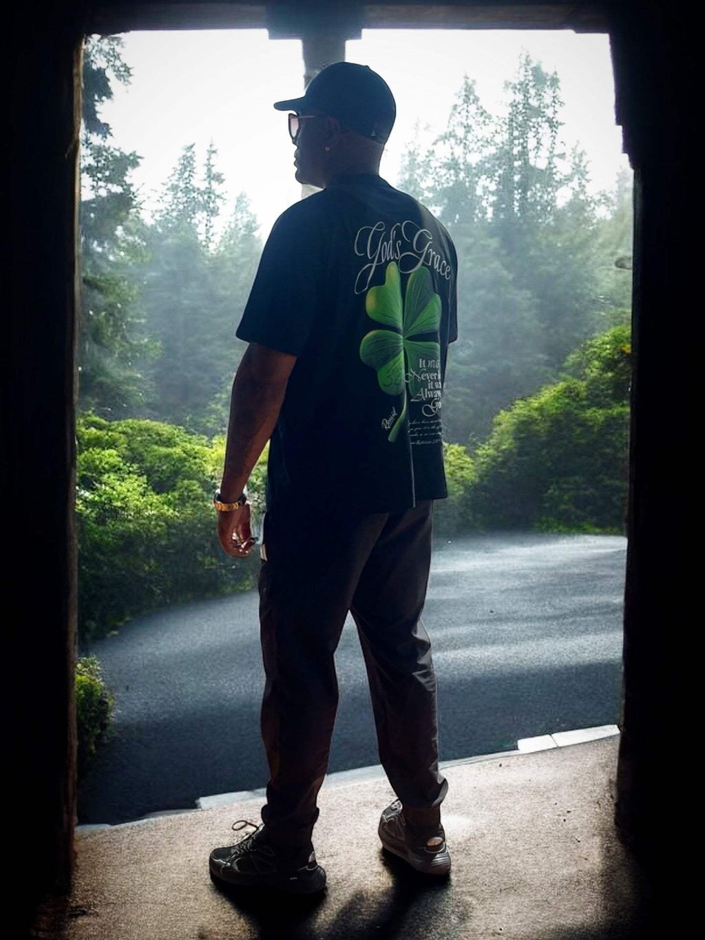Man wearing black God's Grace Oversized Shirt with clover design standing outdoors in nature