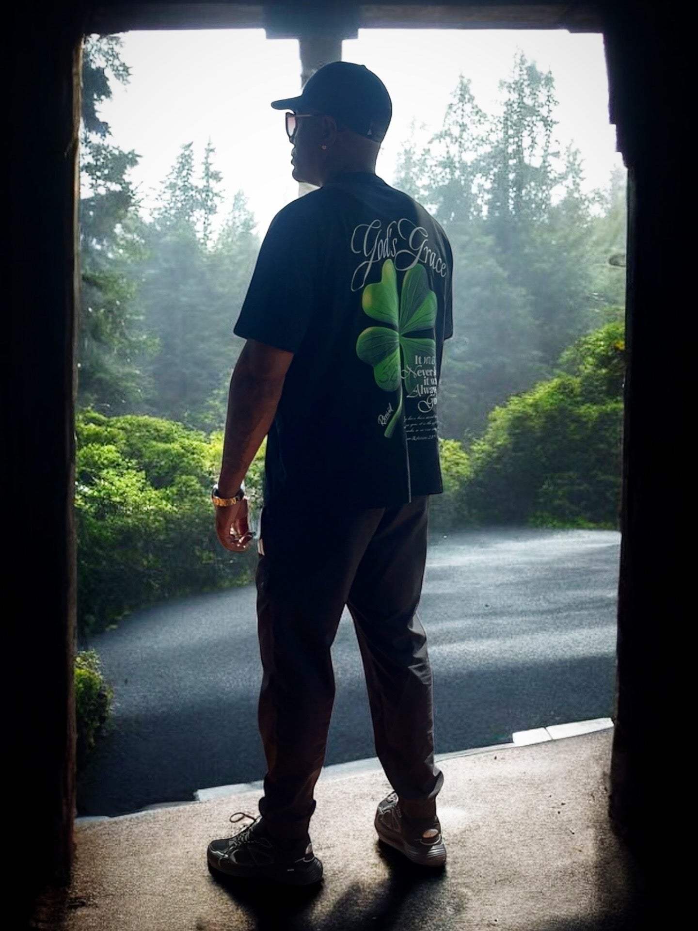 Man wearing black God's Grace Oversized Shirt with clover design standing outdoors in nature